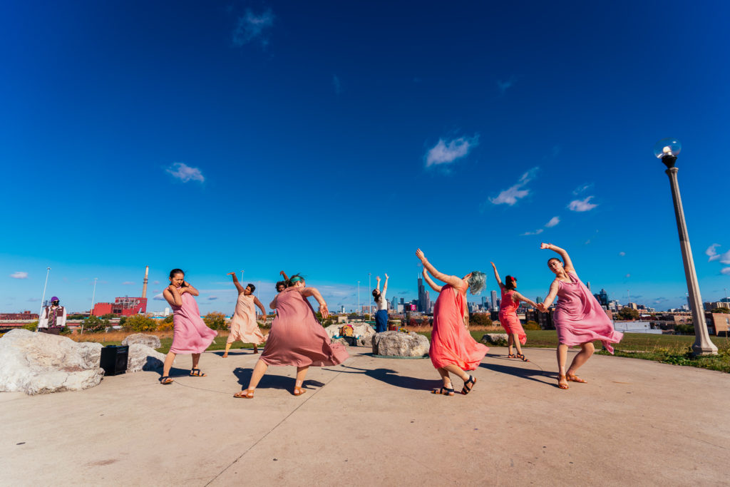 a group of twirling people in a circle wearing flowy orange and pink dresses under a blue sky