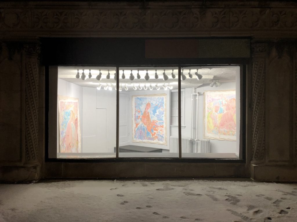 pictured from the windows of the art gallery looking in, a bright white space has multiple large multi-colored paintings